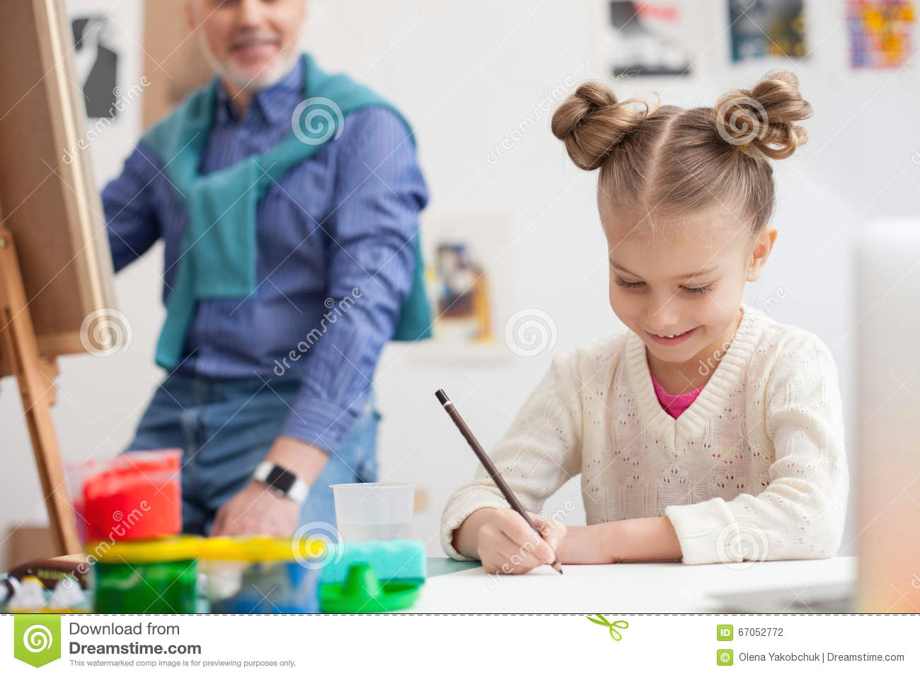 portrait of cute small girl drawing the picture with inspiration she is sitting at the desk and smiling her grandfather is painting and looking at her